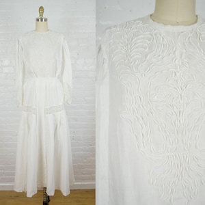May include: A white vintage dress with long sleeves and a floor-length skirt. The bodice and sleeves feature intricate embroidered floral patterns. The dress is displayed on a mannequin against a white brick wall.