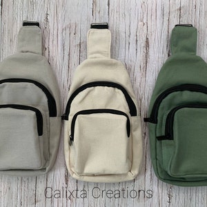 May include: Three canvas sling bags in neutral colors: light gray, cream, and olive green. Each bag features a black zipper, a front pocket, and an adjustable strap. The bags are displayed against a light wood-grain background. The text "Calixta Creations" is visible.