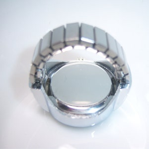 Puede incluir: Reloj de pulsera plateado con esfera redonda y correa de metal. El reloj se muestra desde la parte posterior, revelando la caja de metal.