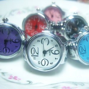 Puede incluir: Seis relojes de anillo plateados con diferentes caras de colores. Las caras son p&uacute;rpura, roja, negra, azul y dos blancas.