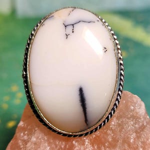 May include: Oval-shaped white gemstone ring with black veining set in a silver-colored metal band. The ring is displayed on a pink, textured surface, with a blurred green background.