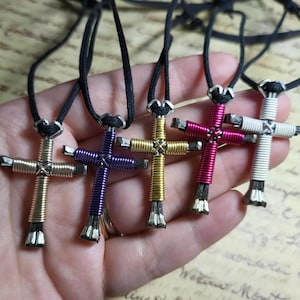May include: A collection of handmade cross necklaces in various colors, including silver, purple, gold, pink, and white. Each cross is crafted with wire and features a black cord for wearing. The crosses are held in a hand.
