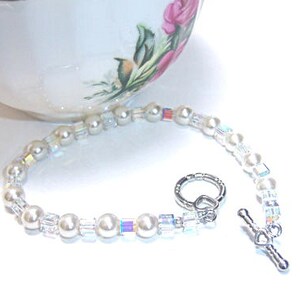 K&ouml;nnte beinhalten: Ein zartes Armband mit wei&szlig;en Kunstperlen und schillernden Kristallperlen. Das Armband hat einen silberfarbenen Knebelverschluss. Eine wei&szlig;e Teetasse mit floralem Design befindet sich im Hintergrund.