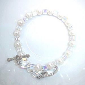 K&ouml;nnte beinhalten: Ein zartes Armband mit einer Mischung aus wei&szlig;en Perlen und schillernden Kristallperlen. Das Armband wird mit einem silberfarbenen Verschluss befestigt. Der Schmuck wird auf einer wei&szlig;en Oberfl&auml;che pr&auml;sentiert.