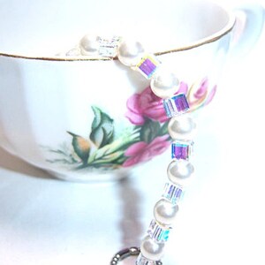 K&ouml;nnte beinhalten: Eine wei&szlig;e Teetasse mit goldenem Rand und Blumendesign, die ein Perlenarmband h&auml;lt. Das Armband besteht aus wei&szlig;en Perlen und schillernden, quadratischen Kristallen. Die Tasse ist mit rosa Blumen und gr&uuml;nen Bl&auml;ttern verziert.