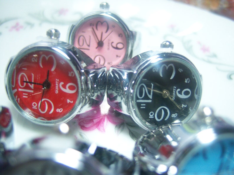 Puede incluir: Tres relojes de pulsera plateados con diferentes caras de colores. El primer reloj tiene una cara roja con n&uacute;meros y manecillas negros. El segundo reloj tiene una cara rosa con n&uacute;meros y manecillas negros. El tercer reloj tiene una cara negra con n&uacute;meros y manecillas blancos.