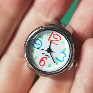 Puede incluir: Un reloj de anillo plateado con una esfera blanca y n&uacute;meros coloridos. La esfera del reloj dice "QUARTZ" y tiene agujas negras.