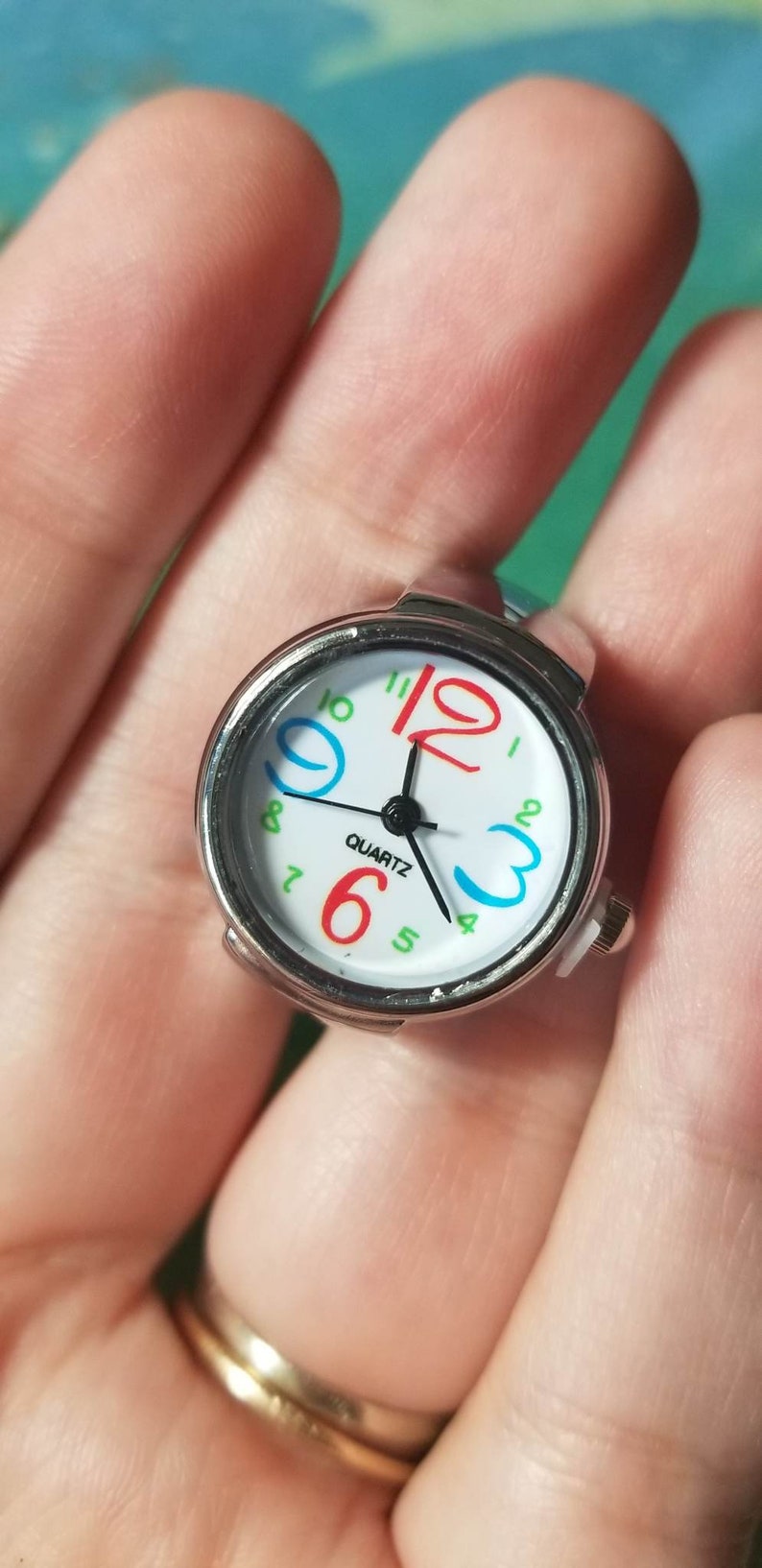 Puede incluir: Un reloj de anillo plateado con una esfera blanca y n&uacute;meros coloridos. La esfera del reloj dice "QUARTZ" y tiene agujas negras.