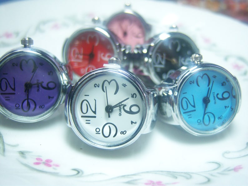 Puede incluir: Seis relojes de anillo plateados con diferentes caras de colores. Las caras son p&uacute;rpura, roja, negra, azul y dos blancas.