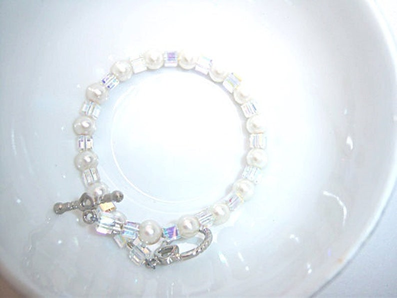 K&ouml;nnte beinhalten: Ein zartes Armband mit einer Mischung aus wei&szlig;en Perlen und schillernden Kristallperlen. Das Armband wird mit einem silberfarbenen Verschluss befestigt. Der Schmuck wird auf einer wei&szlig;en Oberfl&auml;che pr&auml;sentiert.