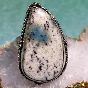 May include: A silver ring featuring a large, teardrop-shaped stone. The stone has a white base with black speckles and a patch of blue. The ring's setting has a twisted rope design around the stone's edge.