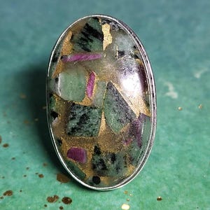May include: Oval-shaped ring featuring a large, polished gemstone. The stone is a mosaic of green, black, gold, and magenta hues, set in a silver-colored metal band. The gemstone's surface has a glossy finish, highlighting the varied colors and textures.