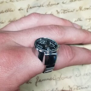 Puede incluir: Un reloj de anillo de plata con una esfera negra y una banda plateada. El reloj est&aacute; en el dedo de una persona.