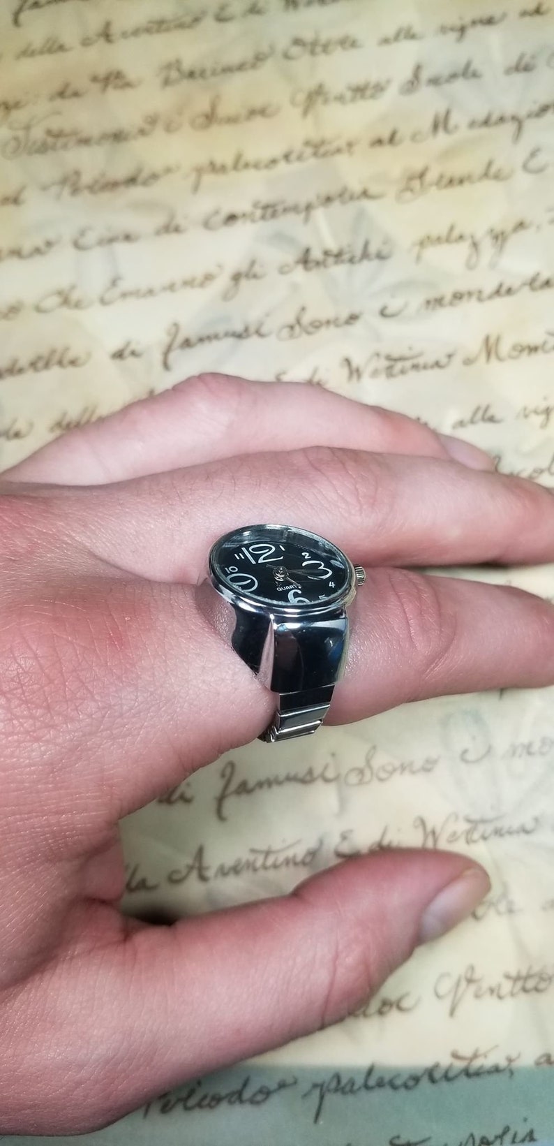 Puede incluir: Un reloj de anillo de plata con una esfera negra y una banda plateada. El reloj est&aacute; en el dedo de una persona.