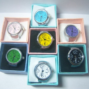 Puede incluir: Surtido de siete relojes de anillo en cajas individuales. Los relojes tienen esferas de diferentes colores: azul, rosa, amarillo, verde, morado, negro y blanco.