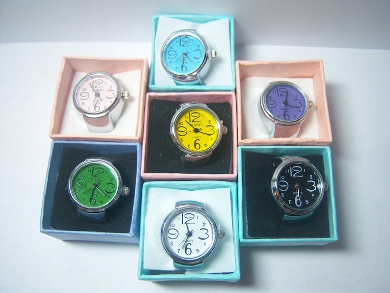 Puede incluir: Surtido de siete relojes de anillo en cajas individuales. Los relojes tienen esferas de diferentes colores: azul, rosa, amarillo, verde, morado, negro y blanco.