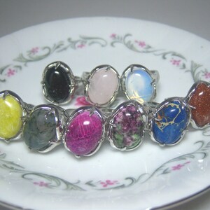 May include: A collection of nine oval-shaped rings featuring various polished gemstones set in silver-toned metal prongs. The stones include yellow, black, light pink, iridescent white, dark green, magenta, mottled green and pink, blue, and gold-flecked brown.
