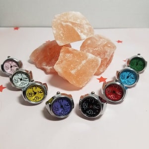 Puede incluir: Surtido de ocho relojes de pulsera coloridos con n&uacute;meros negros en la esfera. Los relojes est&aacute;n en bandas plateadas y est&aacute;n dispuestos en un c&iacute;rculo sobre una superficie blanca. Rocas de sal rosa del Himalaya est&aacute;n en el fondo.