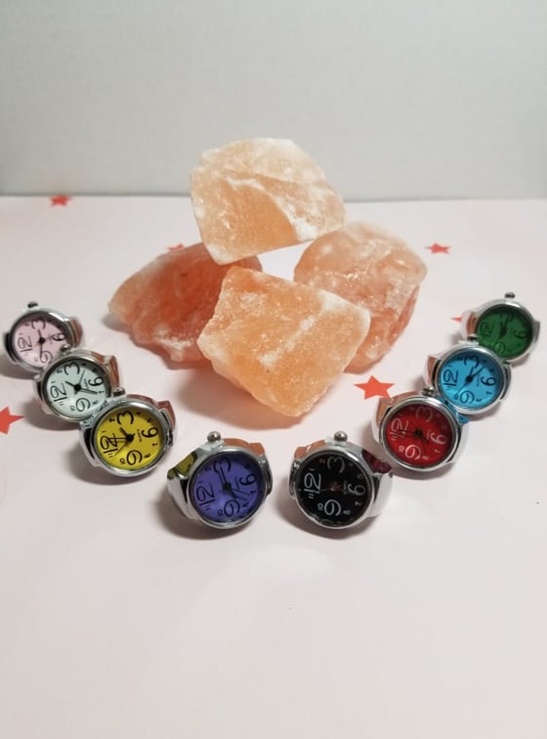 Puede incluir: Surtido de ocho relojes de pulsera coloridos con n&uacute;meros negros en la esfera. Los relojes est&aacute;n en bandas plateadas y est&aacute;n dispuestos en un c&iacute;rculo sobre una superficie blanca. Rocas de sal rosa del Himalaya est&aacute;n en el fondo.