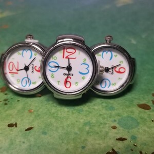 Puede incluir: Tres relojes de pulsera plateados con esferas blancas y n&uacute;meros de colores. Los relojes est&aacute;n conectados entre s&iacute; y tienen un movimiento de cuarzo.