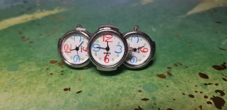 Puede incluir: Tres relojes de pulsera plateados con esferas blancas y n&uacute;meros de colores. Los relojes est&aacute;n conectados entre s&iacute; y tienen un movimiento de cuarzo.