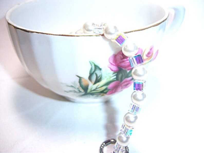 K&ouml;nnte beinhalten: Eine wei&szlig;e Teetasse mit goldenem Rand und Blumendesign, die ein Perlenarmband h&auml;lt. Das Armband besteht aus wei&szlig;en Perlen und schillernden, quadratischen Kristallen. Die Tasse ist mit rosa Blumen und gr&uuml;nen Bl&auml;ttern verziert.