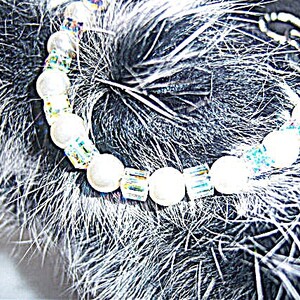 K&ouml;nnte beinhalten: Ein Armband mit wei&szlig;en, runden Perlen und schillernden, rechteckigen Perlen. Das Armband ist vor einem strukturierten, dunklen Hintergrund platziert, der die Details hervorhebt.