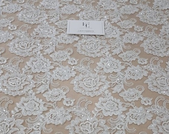 May include: White floral lace fabric with intricate embroidered flower and leaf patterns. The lace features a scalloped edge and is embellished with small, sparkling sequins. The fabric has a delicate, airy appearance, suitable for bridal or formal wear.
