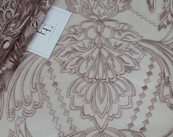 May include: A close-up of a delicate, light brown lace fabric with a floral pattern. The lace has a subtle sheen and a soft, flowing texture.