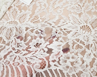 May include: White floral lace fabric featuring an intricate scalloped edge and a delicate mesh background. A small white rectangular tag is attached to the fabric, displaying the black logo and text "LT LACE TO LOVE".