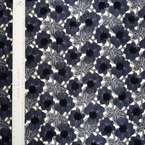 May include: Navy blue floral lace fabric with a repeating pattern of embroidered flowers and leaves. The fabric has a delicate, openwork design, with a ruler along the side. The flowers have a darker center, and the leaves are detailed with fine lines.
