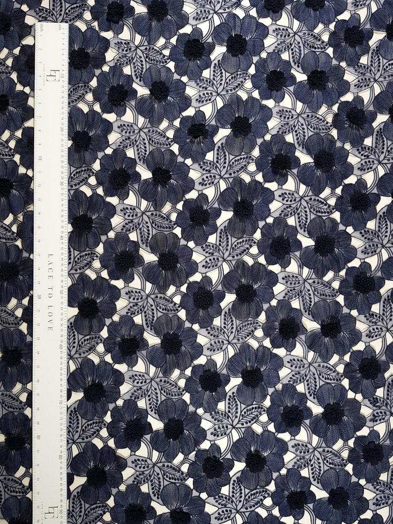 May include: Navy blue floral lace fabric with a repeating pattern of embroidered flowers and leaves. The fabric has a delicate, openwork design, with a ruler along the side. The flowers have a darker center, and the leaves are detailed with fine lines.