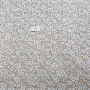 May include: White floral lace fabric with a detailed pattern. The lace features a repeating design of flowers and leaves, creating a textured surface. A small white card with text is visible on the fabric.