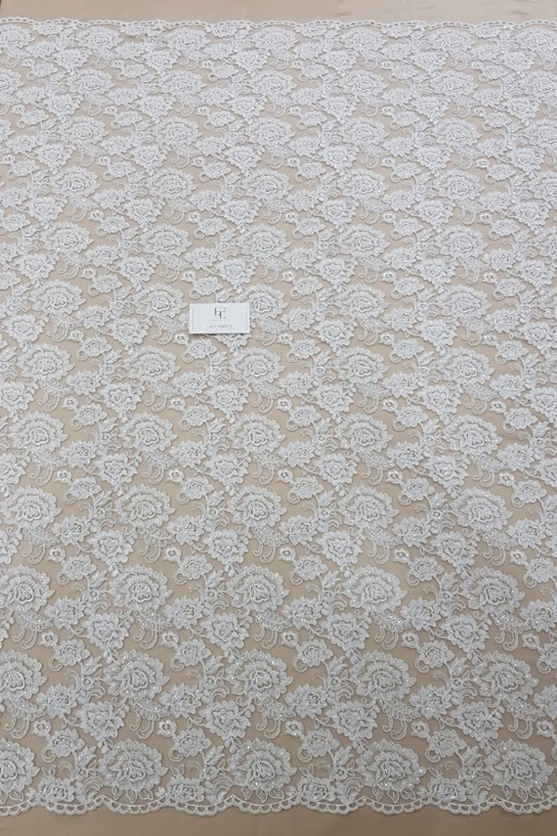 May include: White floral lace fabric with a detailed pattern. The lace features a repeating design of flowers and leaves, creating a textured surface. A small white card with text is visible on the fabric.