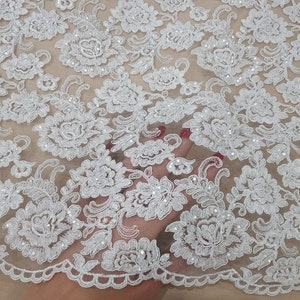 May include: Close-up of white floral lace fabric. The lace features a detailed pattern of flowers and leaves, with small, sparkling embellishments. The fabric has a scalloped edge and is set against a neutral background.
