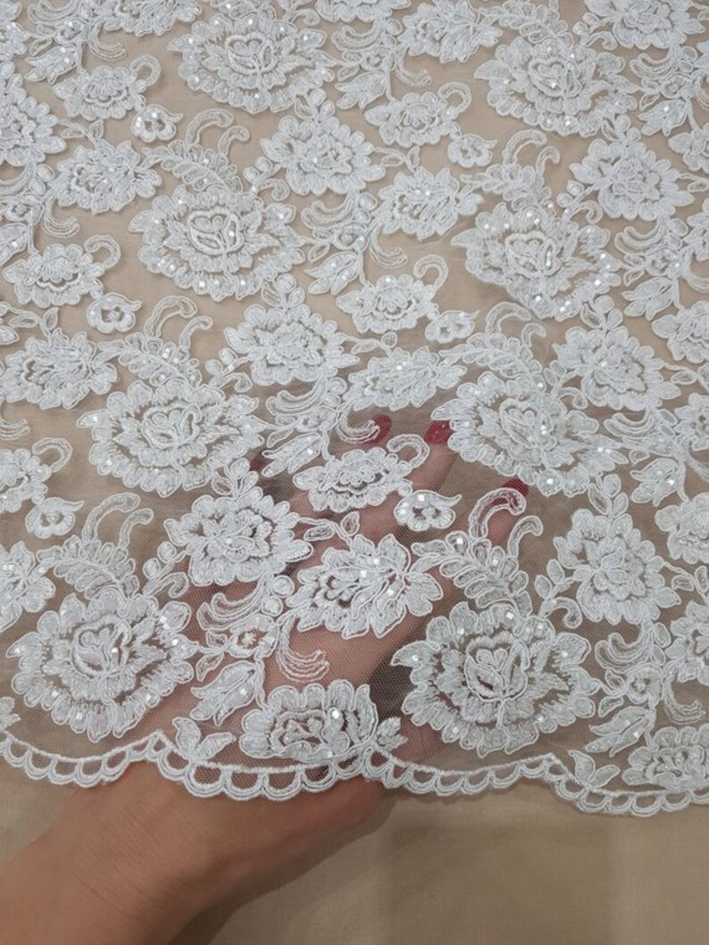 May include: Close-up of white floral lace fabric. The lace features a detailed pattern of flowers and leaves, with small, sparkling embellishments. The fabric has a scalloped edge and is set against a neutral background.