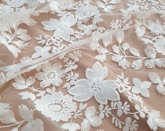 May include: A sheer white fabric featuring intricate floral embroidery with detailed petals and leaves. The white thread work creates a dense, textured pattern of various blossoms and vines layered over a solid, light beige background material.