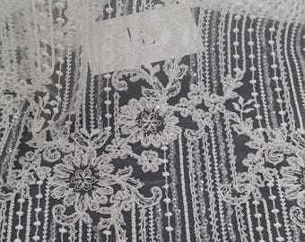 May include: A white lace fabric featuring intricate floral embroidery and vertical beaded fringe detailing. The sheer mesh base is adorned with sparkling sequins and delicate threadwork, creating a layered pattern of flowers and hanging beaded strands.