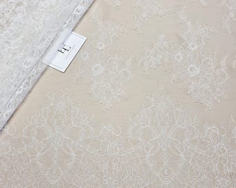 May include: White lace fabric featuring intricate floral and ornate patterns. The lace has a scalloped edge and is partially rolled, displaying the delicate design. A small white card with text is visible.