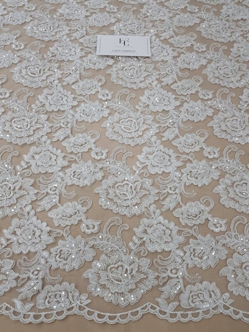 May include: White floral lace fabric with detailed embroidered flower and leaf patterns. The lace has a scalloped edge and is adorned with tiny, shimmering sequins. The fabric has a delicate, airy appearance, suitable for bridal or formal wear.