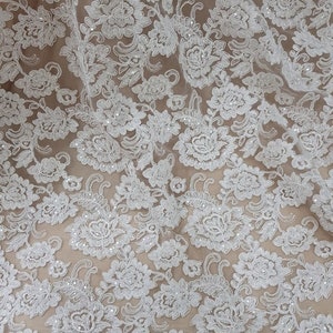 May include: Close-up of white floral lace fabric. The lace features a detailed pattern of flowers and leaves, with a scalloped edge. The delicate design is set against a neutral background, highlighting the intricate details of the lace.