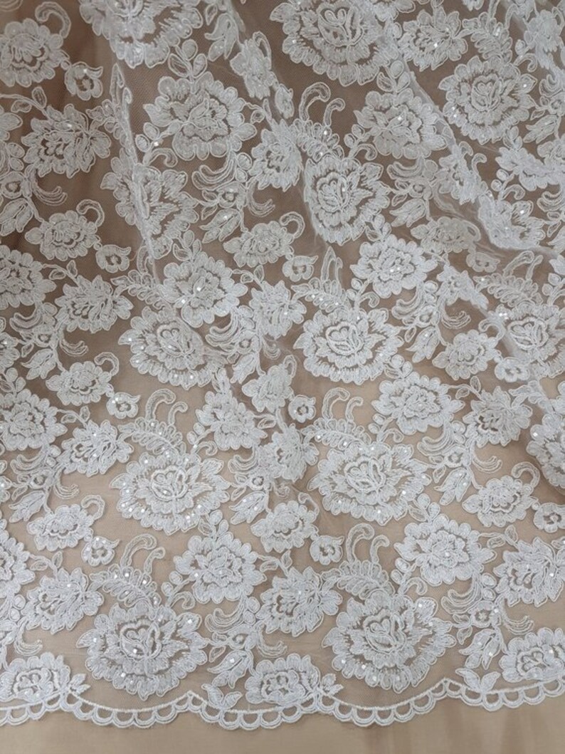 May include: Close-up of white floral lace fabric. The lace features a detailed pattern of flowers and leaves, with a scalloped edge. The delicate design is set against a neutral background, highlighting the intricate details of the lace.