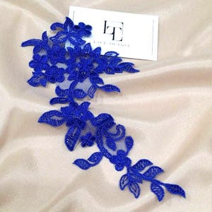 May include: Royal blue floral lace applique with delicate flower and leaf details. The lace is placed on a cream-colored fabric. A white card with the text "LACE TO LOVE" is also present.