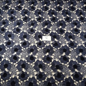May include: Navy blue floral lace fabric. The fabric features a repeating pattern of embroidered flowers with detailed petals and dark centers. The flowers are set against a light background, creating a high-contrast design. The lace is likely used for apparel or decorative purposes.