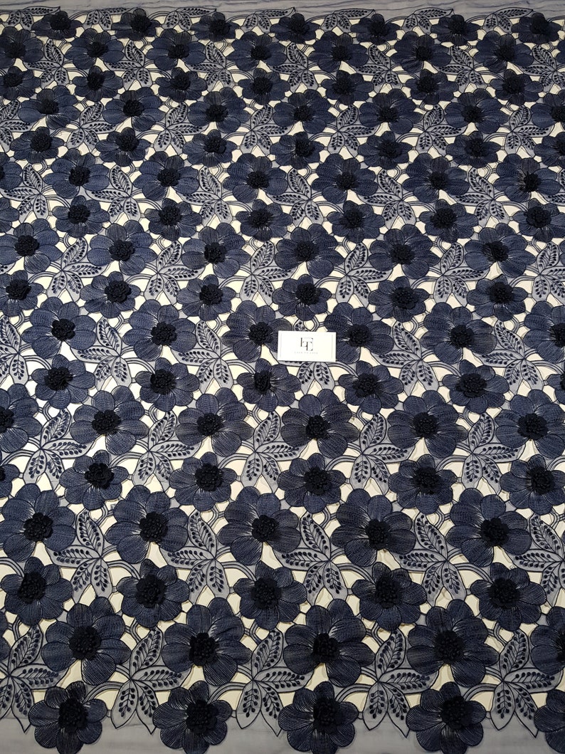 May include: Navy blue floral lace fabric. The fabric features a repeating pattern of embroidered flowers with detailed petals and dark centers. The flowers are set against a light background, creating a high-contrast design. The lace is likely used for apparel or decorative purposes.