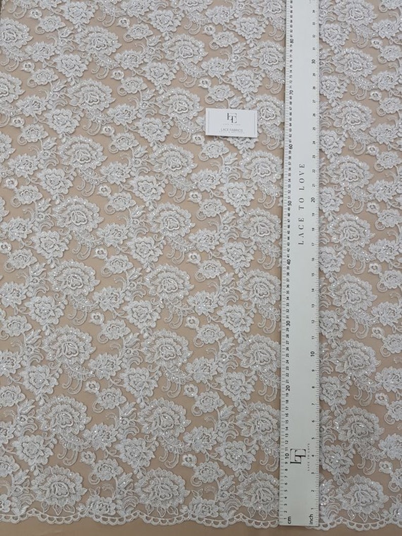 May include: White floral lace fabric featuring a repeating pattern of embroidered flowers and sequins. A ruler with centimetre measurements is visible, along with a tag that reads "Lace Fabrics". The text "Lace to Love" is printed on the ruler.