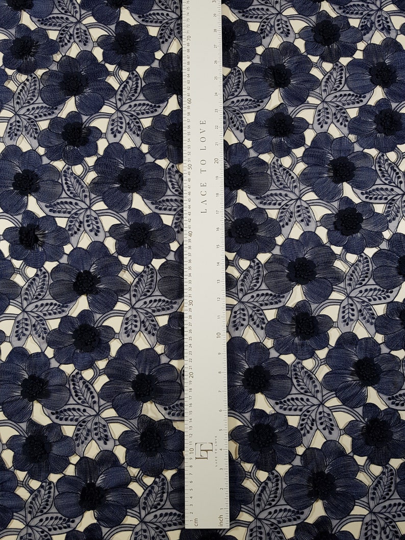 May include: Navy blue floral lace fabric with a repeating pattern of embroidered flowers and leaves. The fabric has a cream-colored background. A ruler with both inch and centimeter measurements is placed vertically on the fabric.