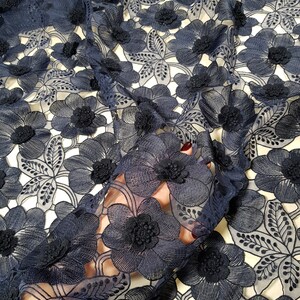 May include: Navy blue floral lace fabric with 3D flower embellishments. The fabric features a repeating pattern of large, detailed flowers and leaves. The flowers have a textured, raised appearance, adding depth and dimension to the design. The lace is semi-sheer, allowing the background to show through.