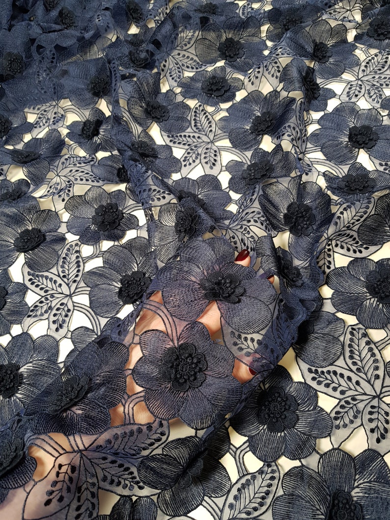 May include: Navy blue floral lace fabric with 3D flower embellishments. The fabric features a repeating pattern of large, detailed flowers and leaves. The flowers have a textured, raised appearance, adding depth and dimension to the design. The lace is semi-sheer, allowing the background to show through.