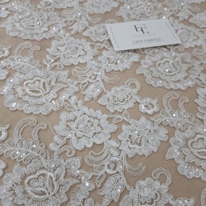 May include: Detailed shot of white floral lace fabric with delicate embroidered patterns. The lace showcases a repeating design of flowers and leaves, adorned with tiny beads. A white card with the text "LACE FABRICS" is on the fabric.